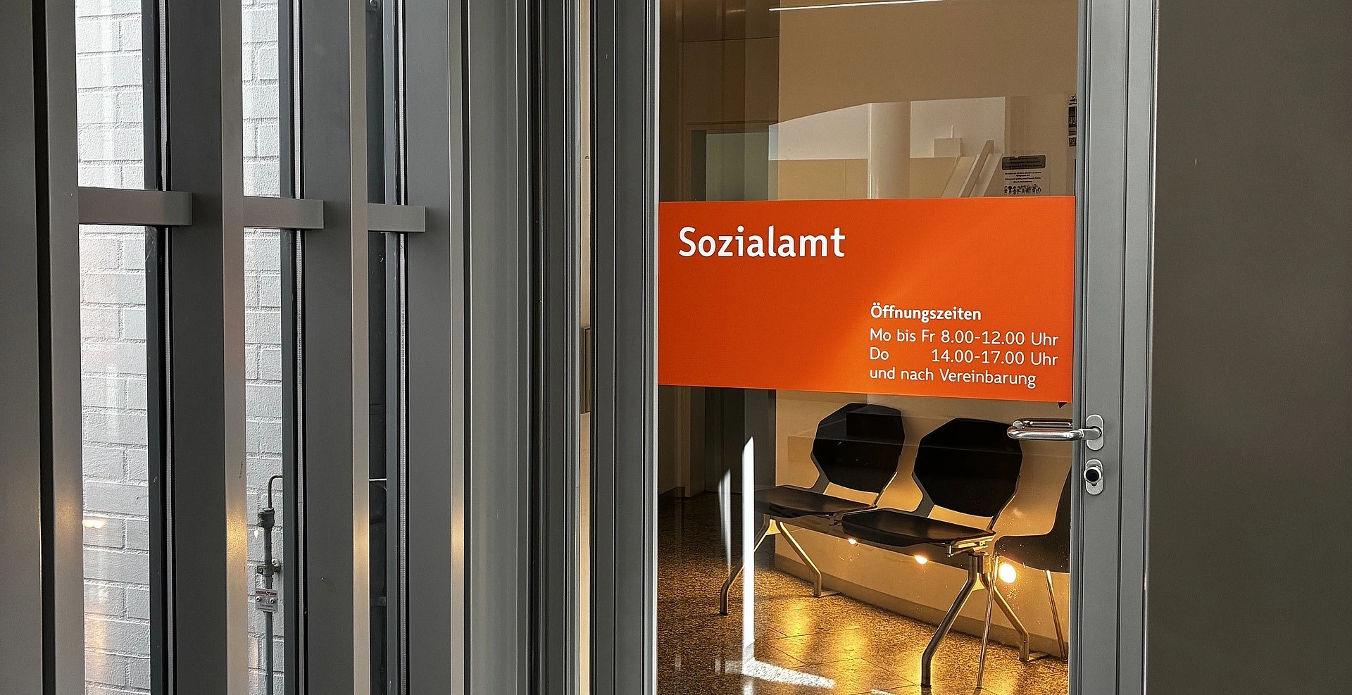 Glastür mit rotem Schild 'Sozialamt' und Öffnungszeiten vor Wartebereich mit Stühlen