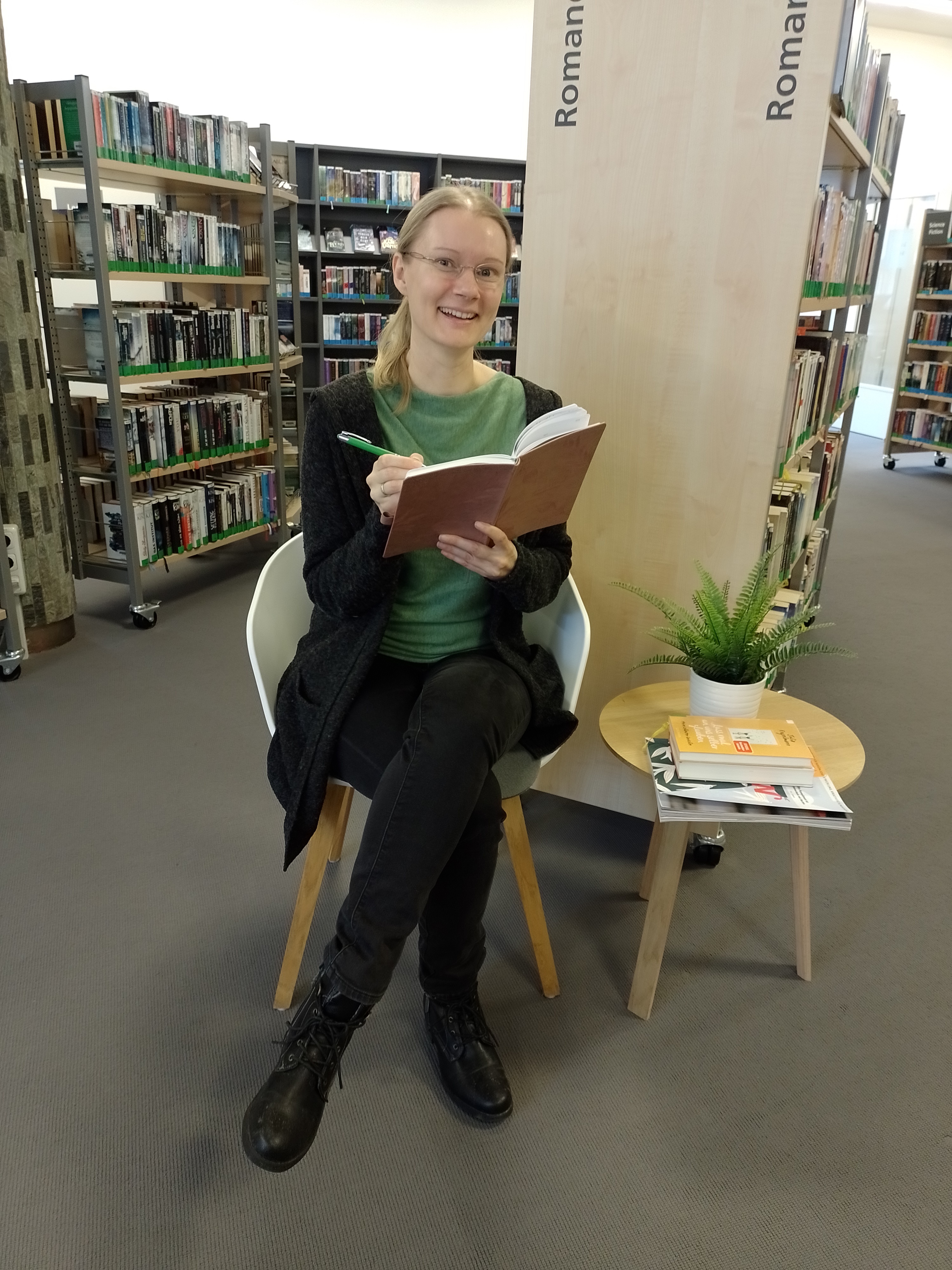Bild vergrößern: Eine Blonde Frau sitzt in der Bibliothek auf einem Stuhl mit Buch und Stift in der Hand